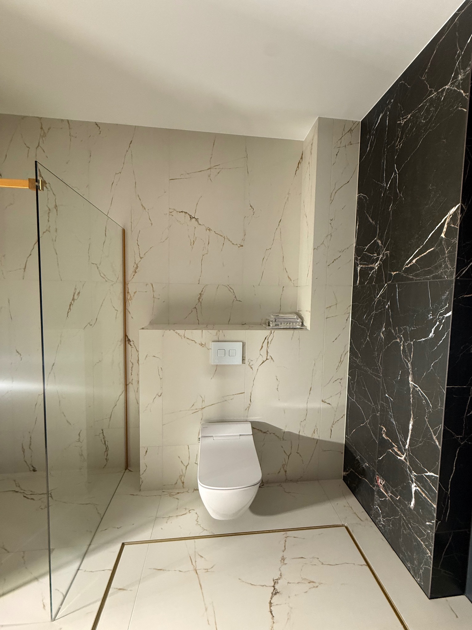 Elegancka łazienka po remoncie: białe i czarne marmurowe płytki, kabina prysznicowa ze złotymi detalami, nowoczesna toaleta. Widok na wykończone wnętrze.