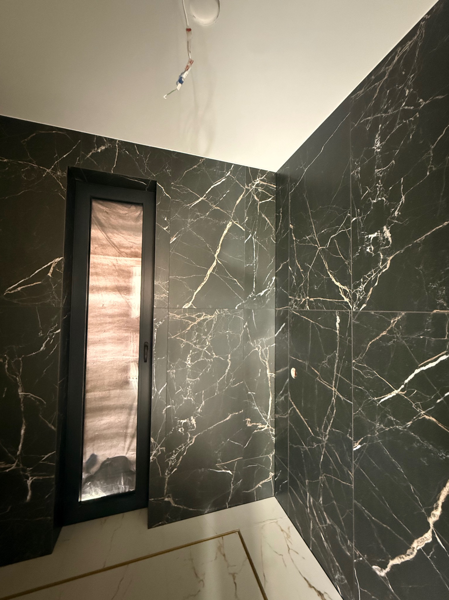 Elegancka łazienka z czarnymi płytkami imitującymi marmur z białymi żyłami, czarna rama drzwi, biało-złota podłoga. Widoczne niedokończone podłączenie oświetlenia.