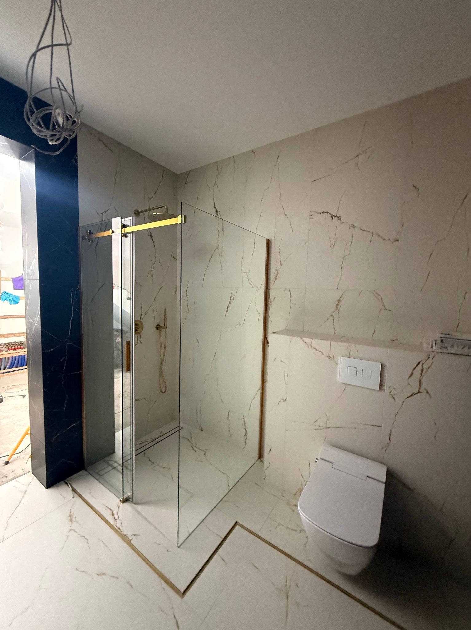 Wykończona łazienka z kabiną prysznicową typu walk-in, białą toaletą i płytkami imitującymi marmur z złotymi akcentami. Widoczne niedokończone oświetlenie.