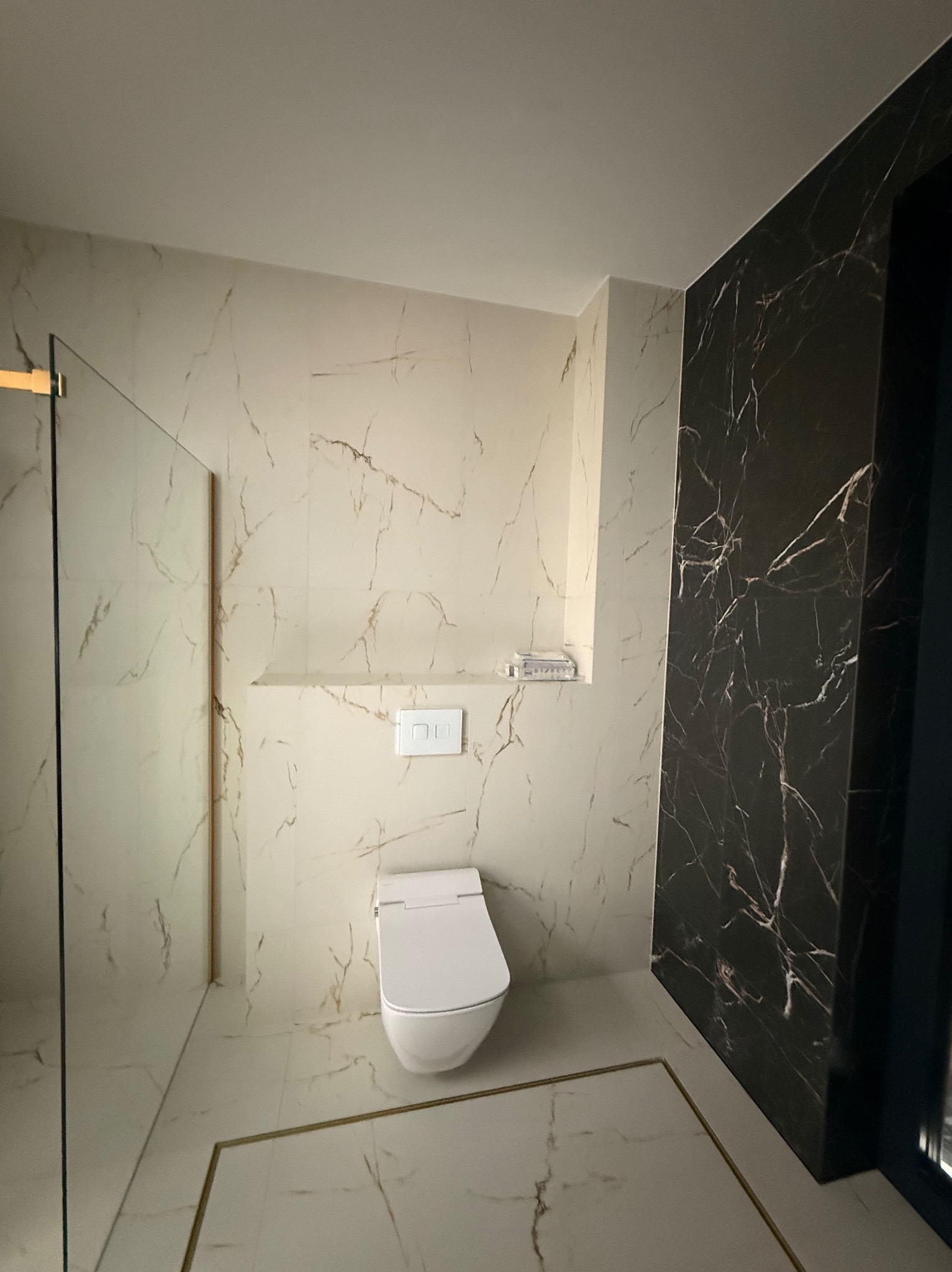 Elegancka łazienka z marmurowymi płytkami w odcieniach bieli i czerni, minimalistyczna toaleta, szklana kabina prysznicowa z złotymi akcentami i wnęka z półką.