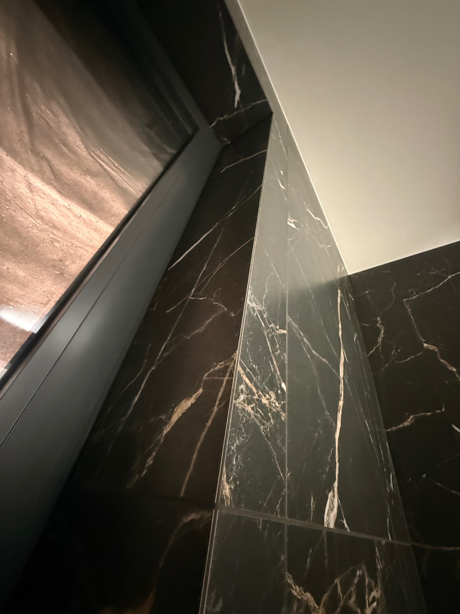 Ciemna, marmurowa ściana z widocznym fragmentem okna i sufitu. Ułożenie płytek tworzy geometryczny wzór. Wykończenie wnętrza w nowoczesnym stylu.