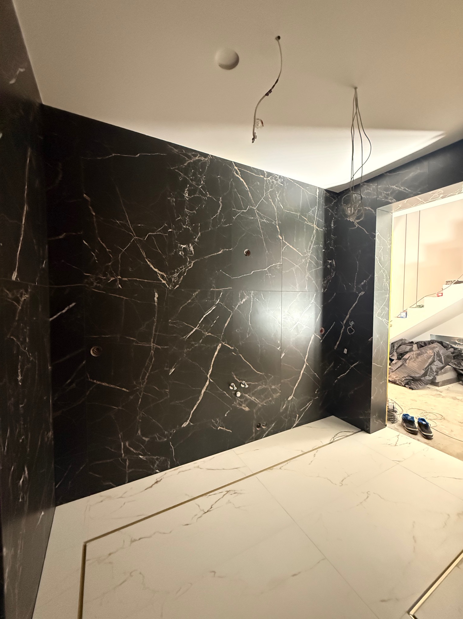 Częściowo wykończona łazienka z czarnymi płytkami imitującymi marmur na ścianach i białymi na podłodze, z widocznymi przewodami elektrycznymi.