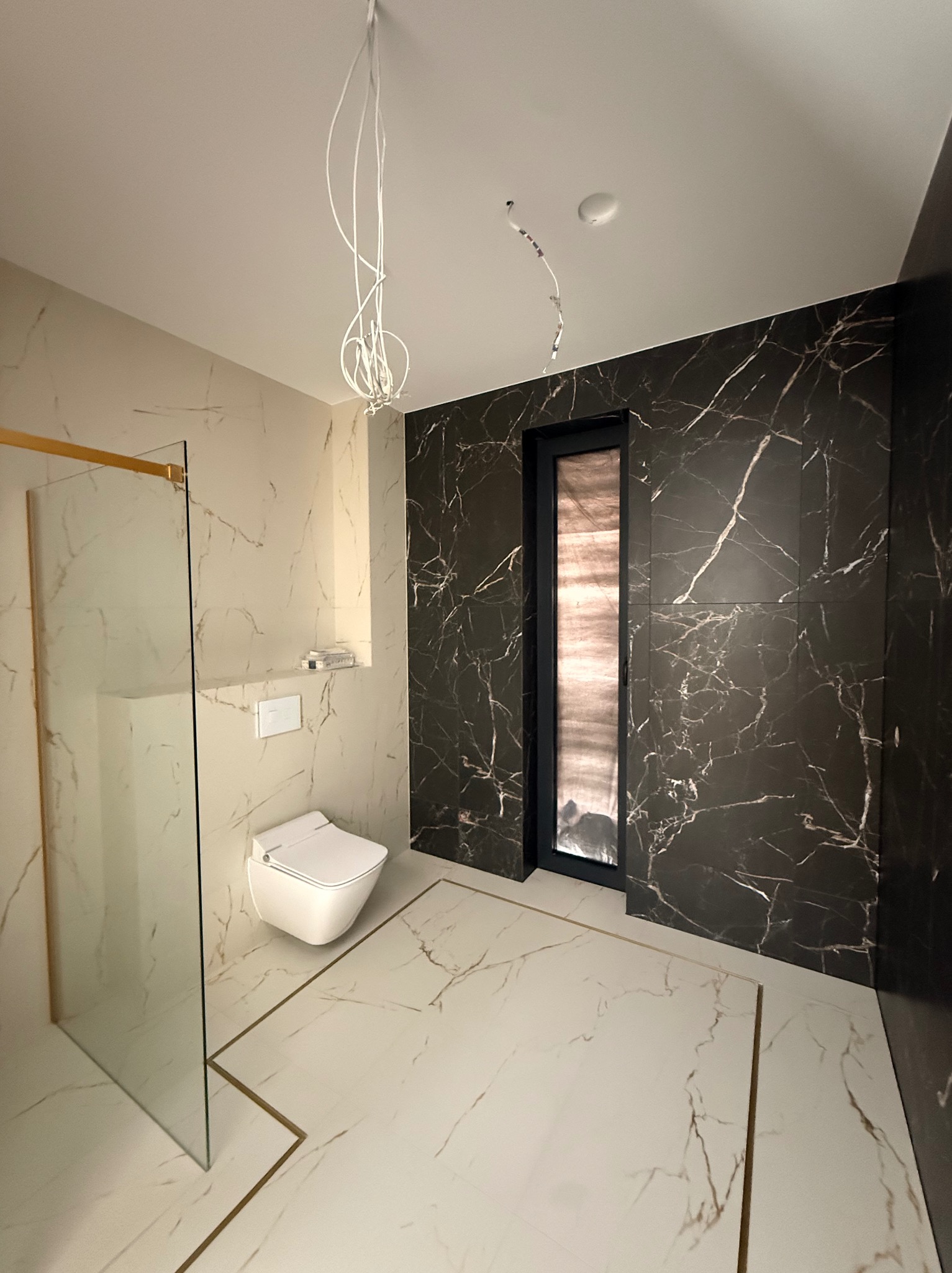 Elegancka łazienka z marmurowymi płytkami w odcieniach bieli i czerni, kabiną prysznicową ze złotymi detalami i podwieszaną toaletą. Widoczne niedokończone podłączenia oświetlenia.