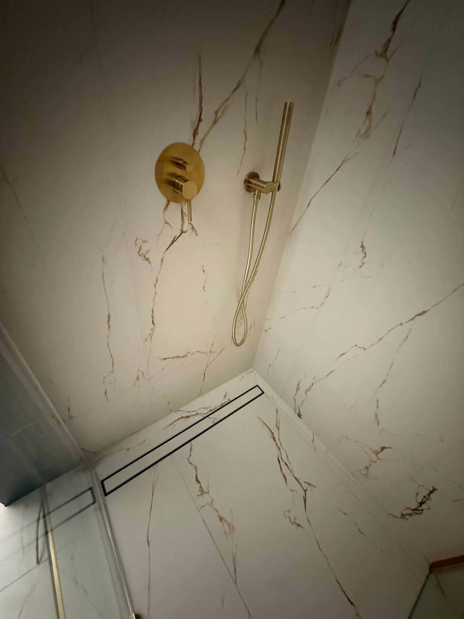 Wykończona kabina prysznicowa z białymi płytkami imitującymi marmur, złotą armaturą i czarnym odpływem liniowym. Widok z dołu, fragment szklanej ściany kabiny.