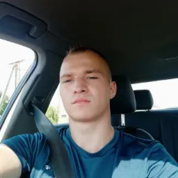 Młody mężczyzna w niebieskiej koszulce siedzi w samochodzie, zapięty pasem bezpieczeństwa, z poważnym wyrazem twarzy, w jasnym świetle słonecznym.