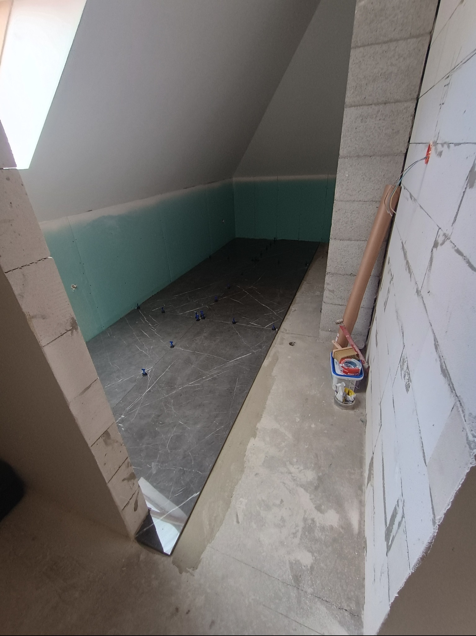 Układanie ciemnych płytek w pomieszczeniu na poddaszu. Widoczne krzyżyki dystansowe, ściany z bloków i płyty gipsowe. Częściowo ukończona podłoga.