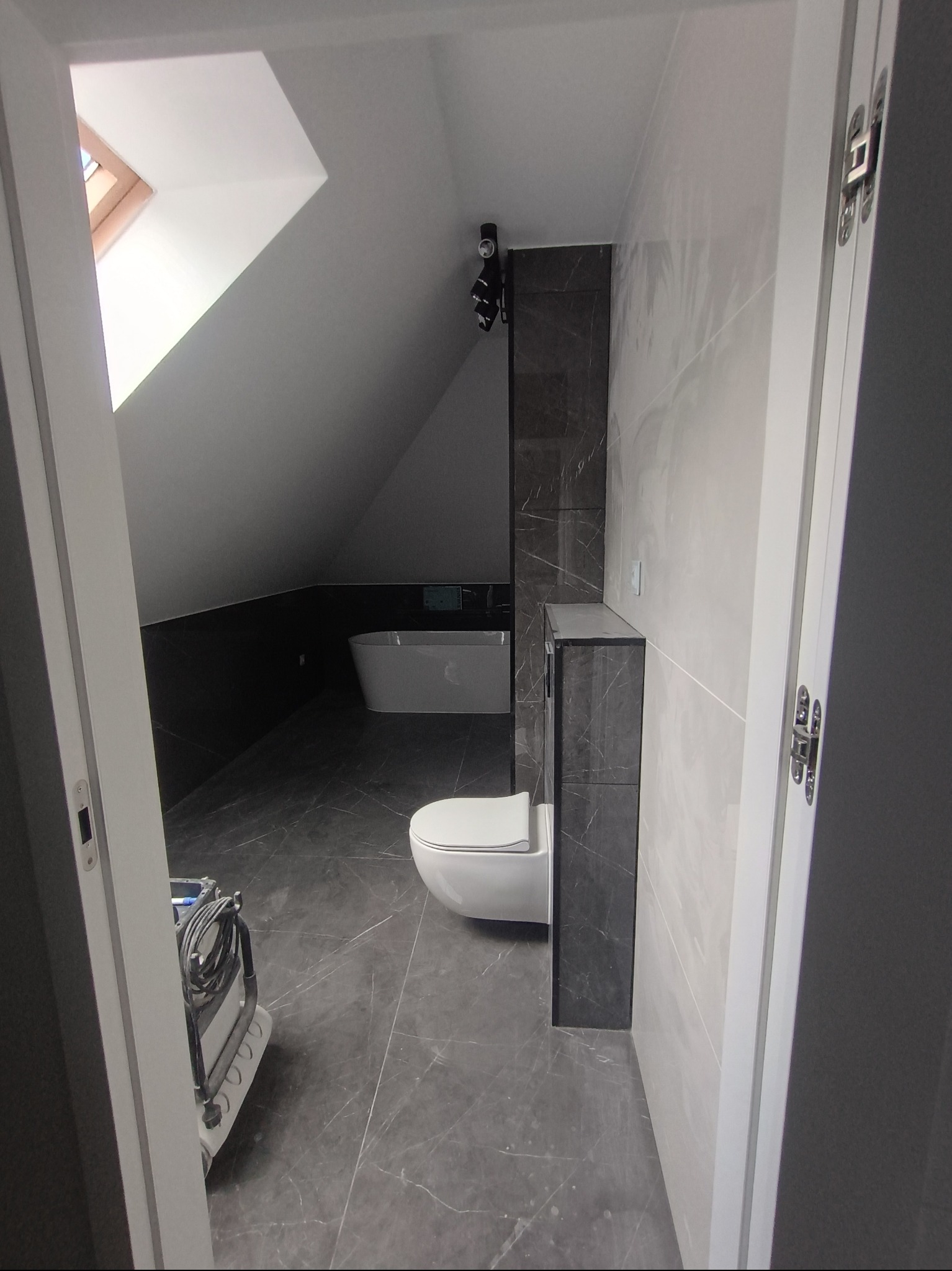 Nowoczesna łazienka na poddaszu z marmurową podłogą i ścianami. Widoczna wanna, toaleta i szafka. Ujęcie przez otwarte drzwi, z widocznym sprzętem do sprzątania.