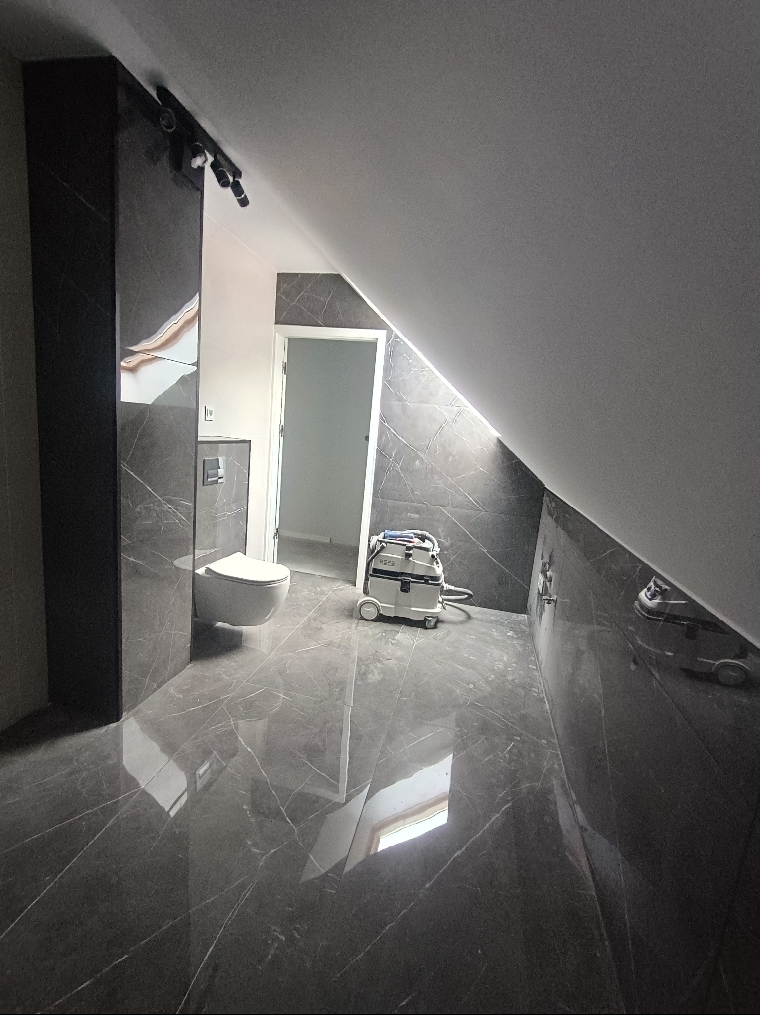 Wykończona łazienka na poddaszu z szarymi płytkami imitującymi marmur, wiszącą toaletą i odkurzaczem przemysłowym. Widoczne oświetlenie punktowe.