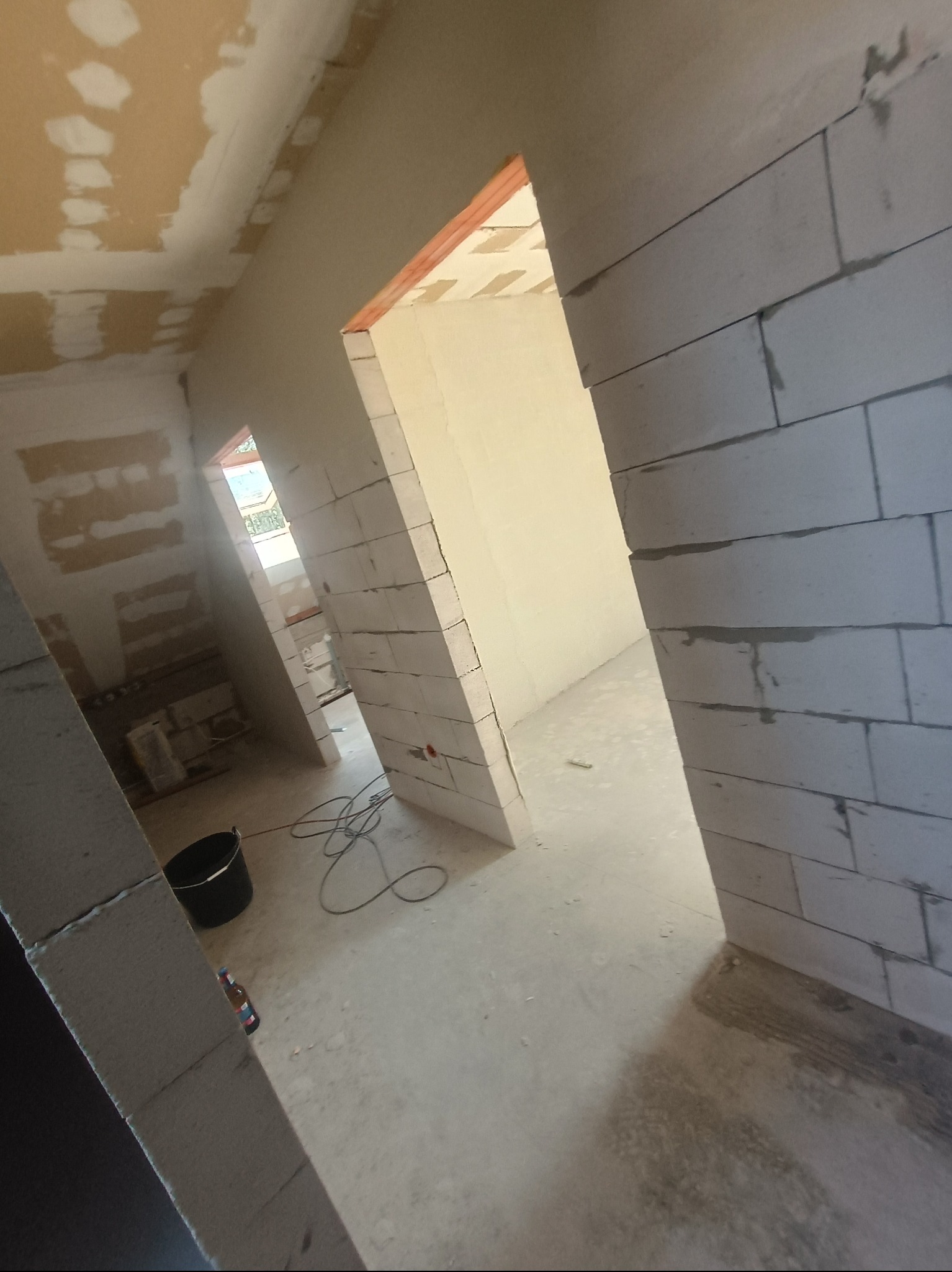 Wnętrze w trakcie budowy: surowe ściany z białych bloczków, widoczne przewody i wiadro. Sufit częściowo wykończony, w tle okno z widokiem na zieleń. Ujęcie z dołu.