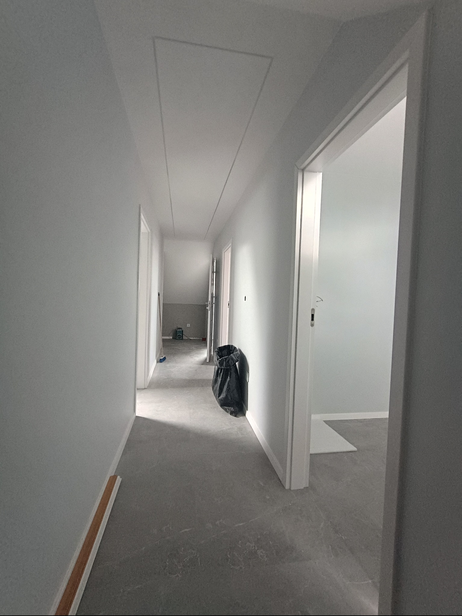 Korytarz w szarościach po remoncie: świeżo pomalowane ściany, szara podłoga, widoczny właz na strych i worek z odpadami. Minimalistyczne wykończenie wnętrza.