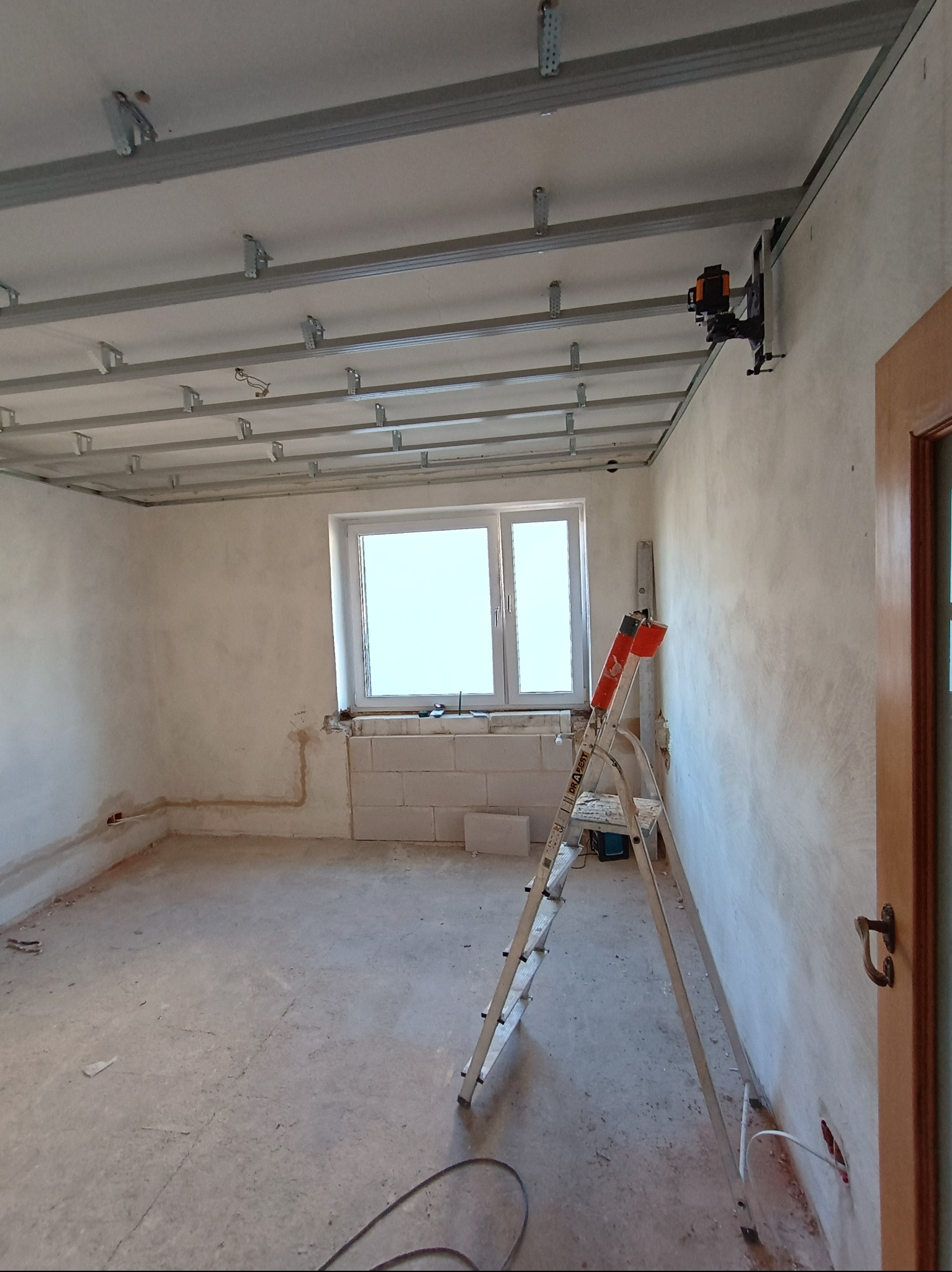 Montaż sufitu podwieszanego z profili metalowych, widoczna drabina i laserowy niwelator. W tle okno i ściany w trakcie prac wykończeniowych.