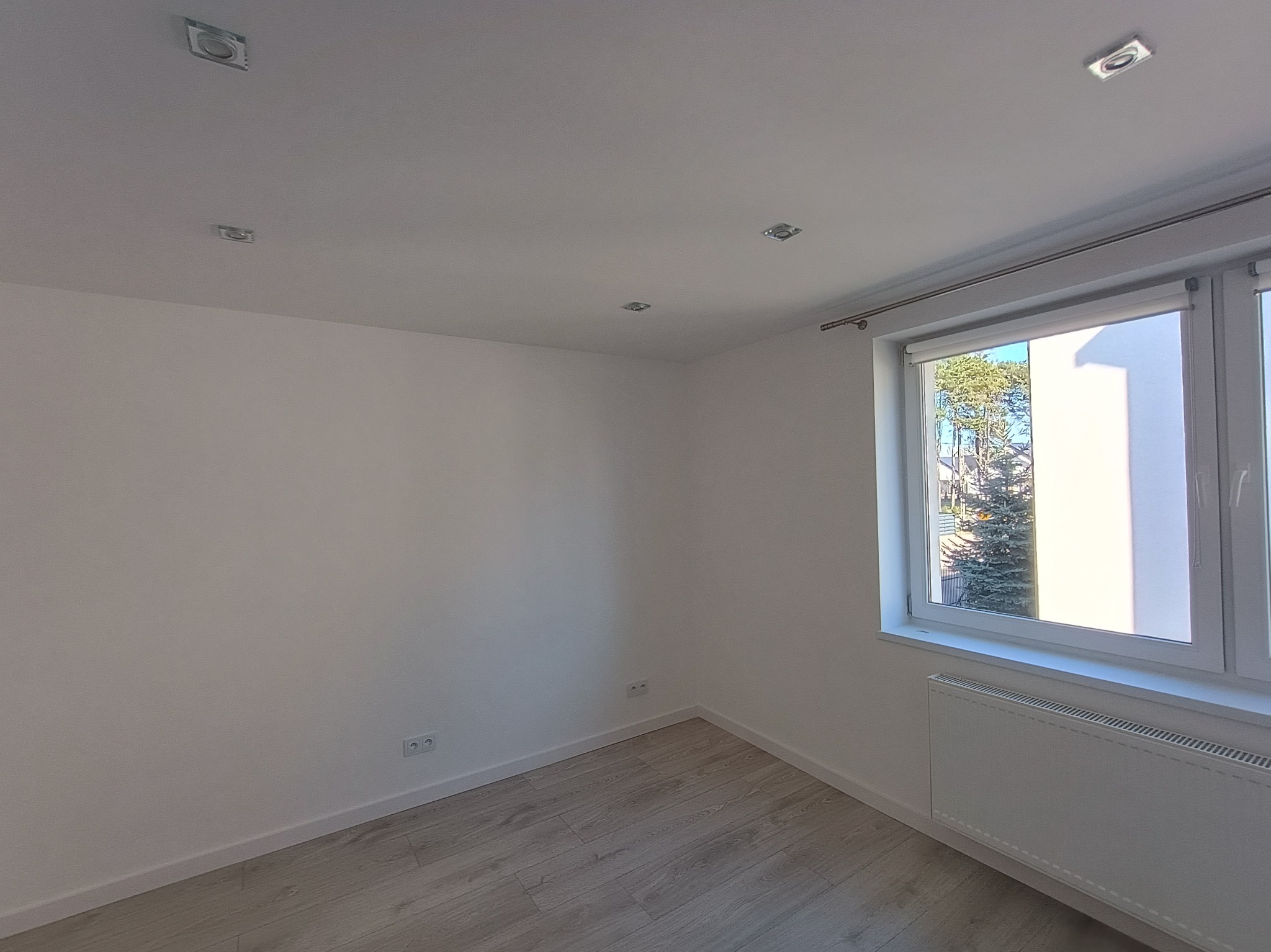 Jasne wnętrze pokoju po remoncie: białe ściany, panele podłogowe w odcieniach szarości, okno z widokiem na zieleń i grzejnik. Minimalistyczne oświetlenie sufitowe.