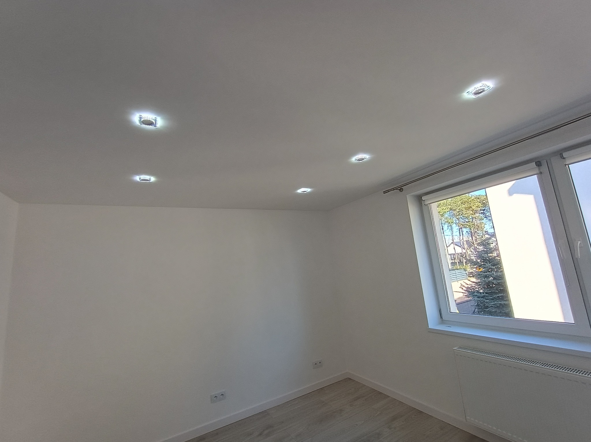 Jasne wnętrze po remoncie: białe ściany, parkiet, okno z widokiem na zieleń i nowoczesne oświetlenie sufitowe. Minimalistyczny design, świeżo wykończone gniazdka.