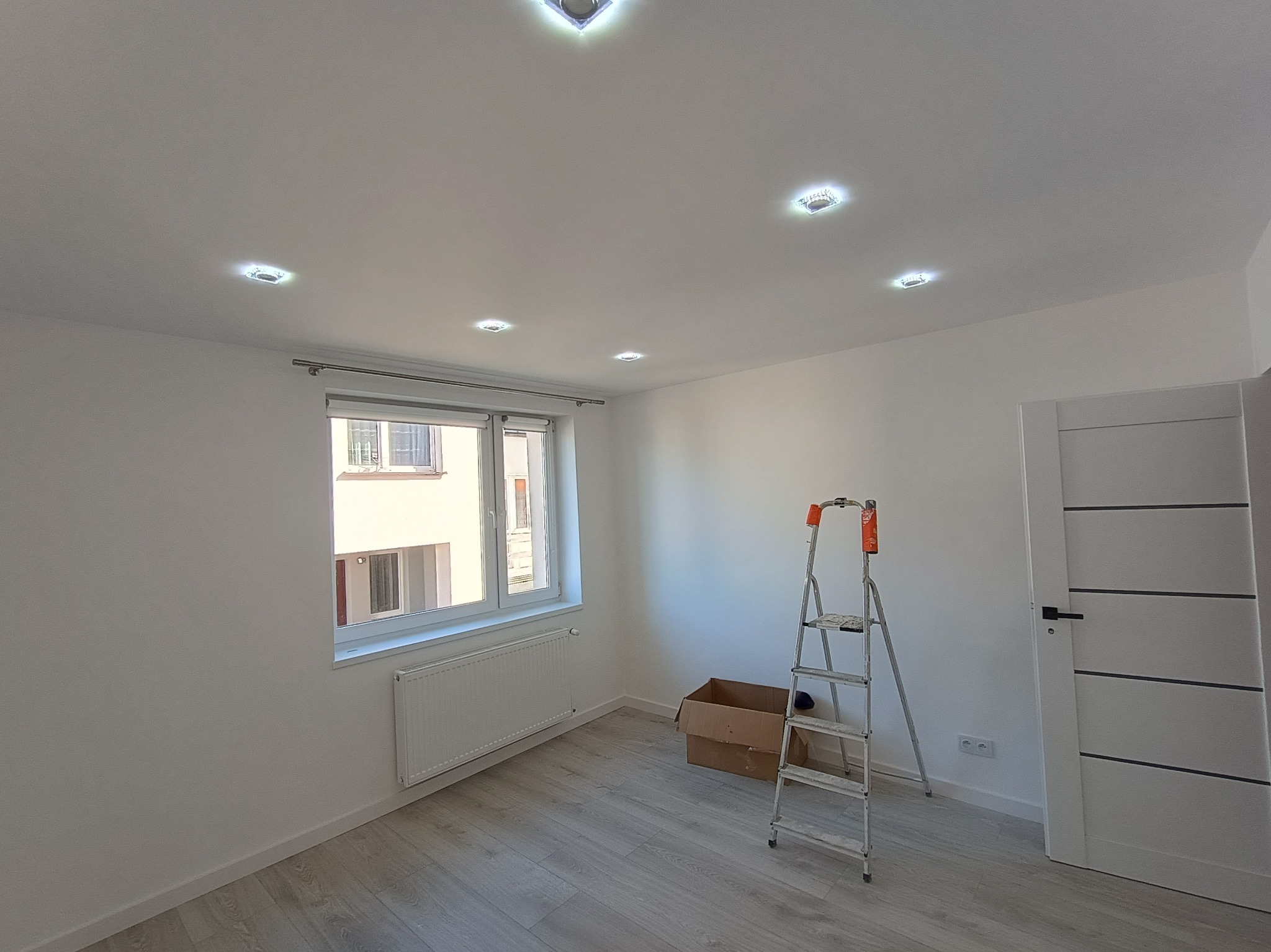 Jasne wnętrze po remoncie: białe ściany, panele podłogowe, okno z widokiem na budynek, drabina malarska i zamontowane oświetlenie sufitowe.