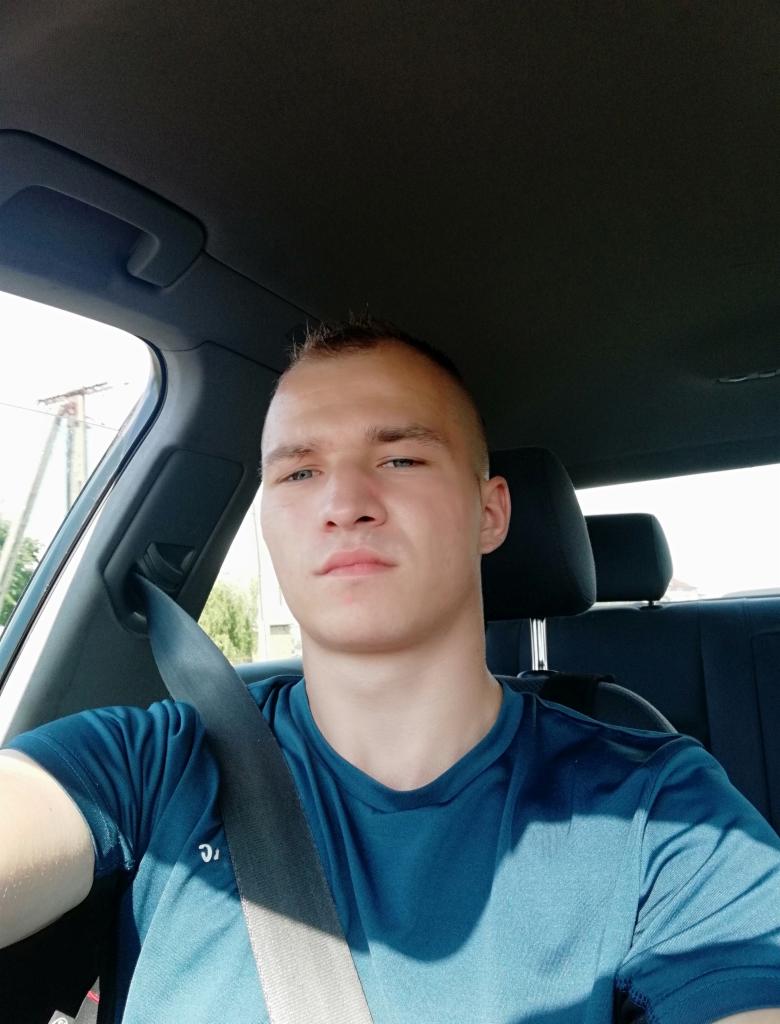 Młody mężczyzna w niebieskiej koszulce siedzi w samochodzie, zapięty pasem bezpieczeństwa, z poważnym wyrazem twarzy, w jasnym świetle słonecznym.