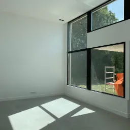 Wnętrze z dużymi oknami z czarnymi ramami, widok na zewnątrz z rusztowaniem i pomarańczową tkaniną, światło słoneczne na szarej podłodze, surowy stan wykończenia.