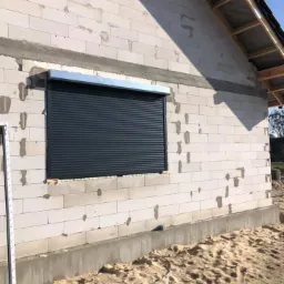 Szara roleta zewnętrzna zamontowana w oknie budynku z białego bloczka, widoczny fragment ramy okiennej po lewej stronie, piasek u podstawy ściany.