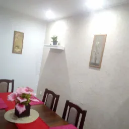 Jasne wnętrze jadalni z drewnianym stołem nakrytym czerwonym bieżnikiem i różowymi podkładkami, na stole dekoracja kwiatowa, na ścianach obrazy i półka z rośliną.