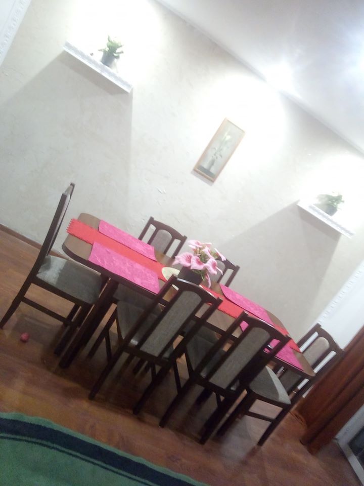 Stół jadalny z ciemnego drewna, nakryty różowymi bieżnikami, otoczony krzesłami z szarym obiciem. Na stole wazon z różowymi kwiatami. W tle jasne ściany z półkami i dekoracją.