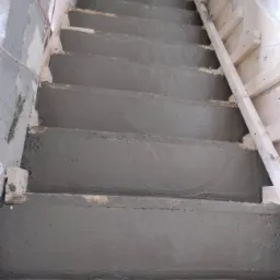 Wewnętrzne schody w trakcie wylewania betonem, widoczne drewniane szalunki po prawej stronie i fragment instalacji elektrycznej po lewej, surowy stan wykończenia.