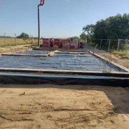 Fundament domu w trakcie budowy, pokryty czarną folią hydroizolacyjną, z wystającymi prętami zbrojeniowymi i stosem cegieł Porotherm w tle, rura do betonu na pierwszym planie.