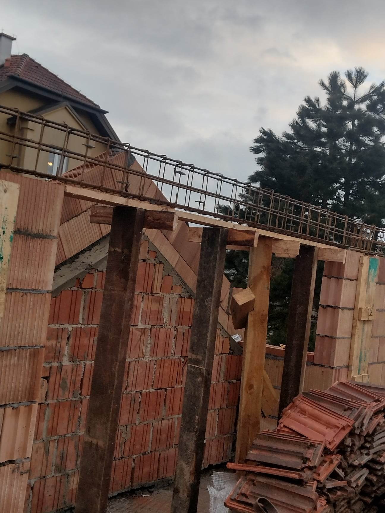 Wznoszenie muru z czerwonej cegły ceramicznej, widoczne drewniane stemple podtrzymujące konstrukcję, w tle fragment dachu sąsiedniego budynku i sosna.