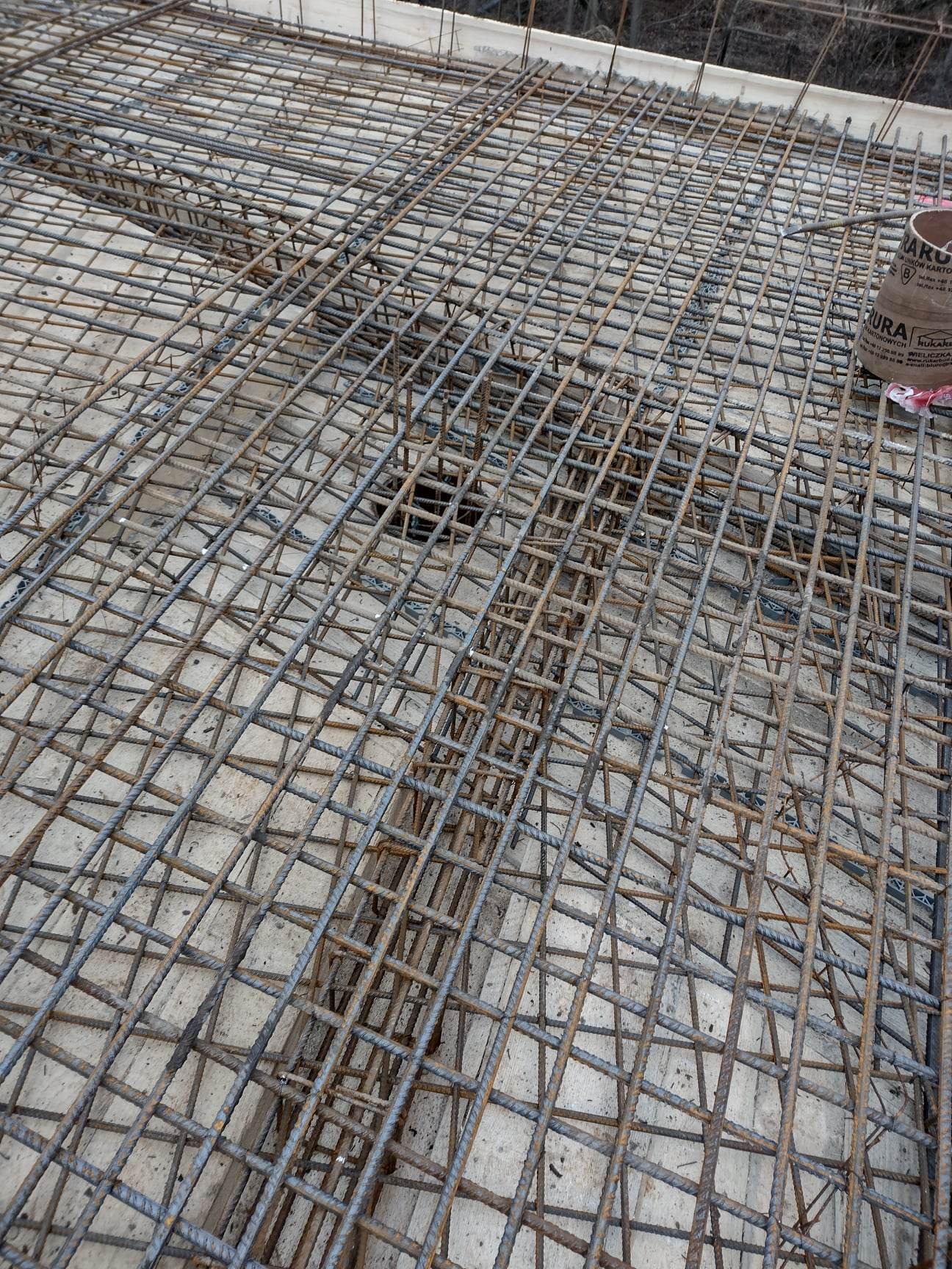 Siatka zbrojeniowa ułożona na betonowym fundamencie, widoczne elementy szalunku i fragment puszki z klejem.