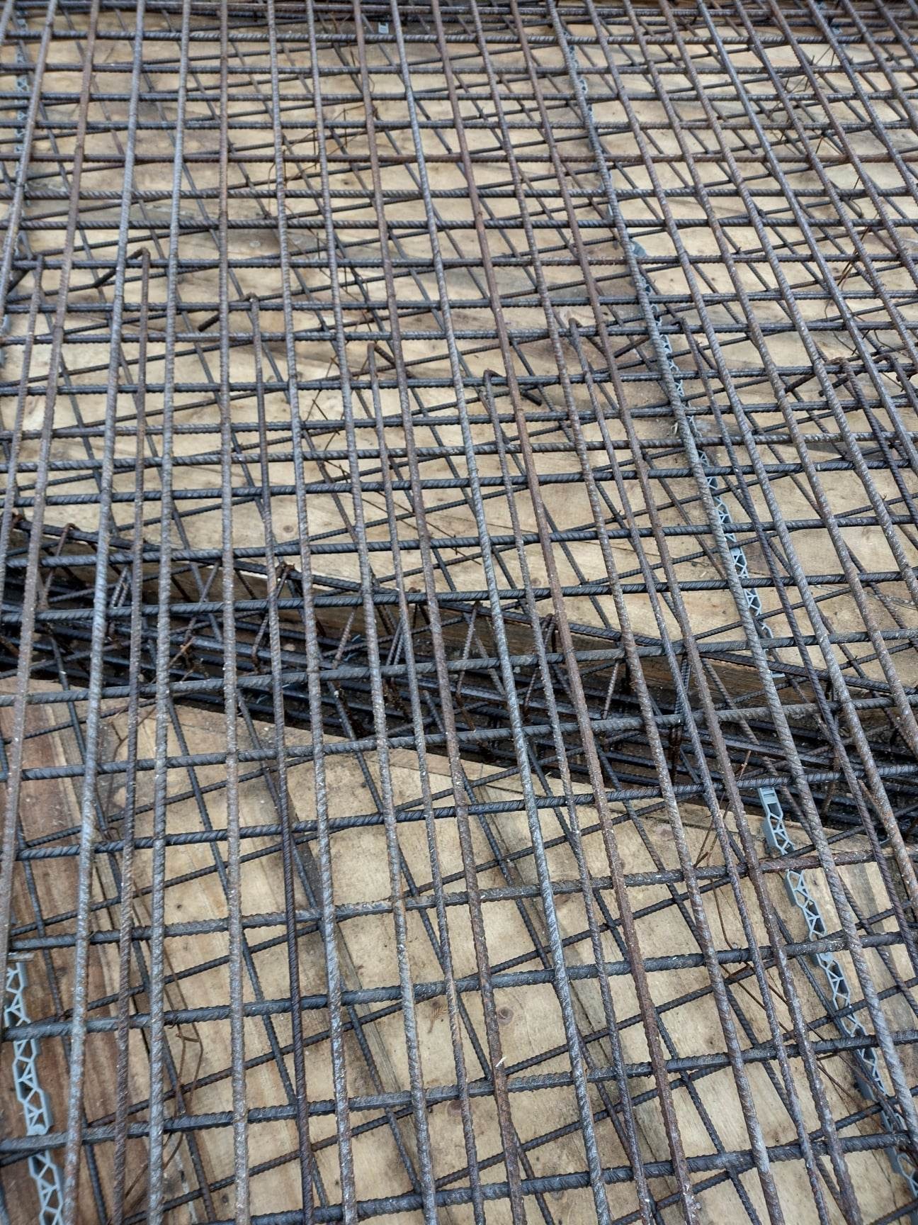 Siatka zbrojeniowa ułożona na deskach szalunkowych, widoczne plastikowe dystanse, przygotowanie do wylewania fundamentów.