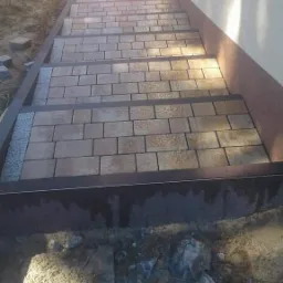 Schody zewnętrzne wyłożone kostką brukową w trakcie budowy, widoczne fragmenty ziemi i gruzu, obrzeża schodów z ciemnego metalu, jasna elewacja budynku w tle.