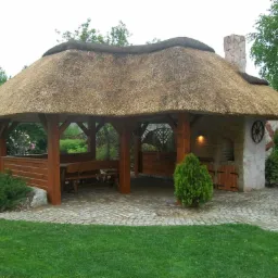 Zadaszona altana ogrodowa z dachem krytym strzechą, drewnianą konstrukcją, murowanym grillem i kamienną posadzką z kostki brukowej, otoczona zielenią.