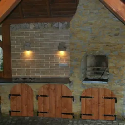 Zadaszona, murowana kuchnia ogrodowa z grillem, blatem roboczym i drewnianymi drzwiczkami, oświetlona dwoma kamiennymi kinkietami.