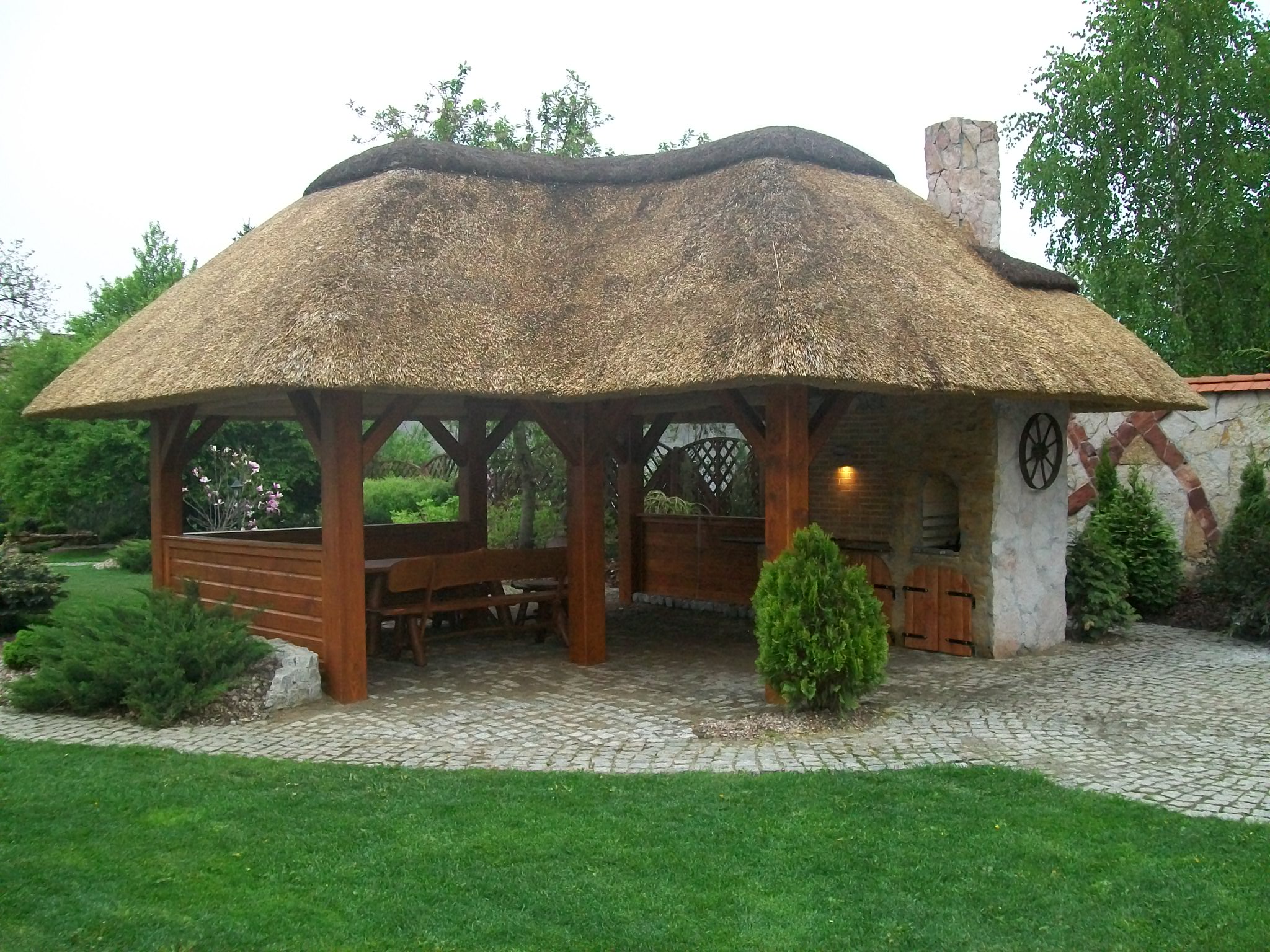 Zadaszona altana ogrodowa z dachem krytym strzechą, drewnianą konstrukcją, murowanym grillem i kamienną posadzką z kostki brukowej, otoczona zielenią.
