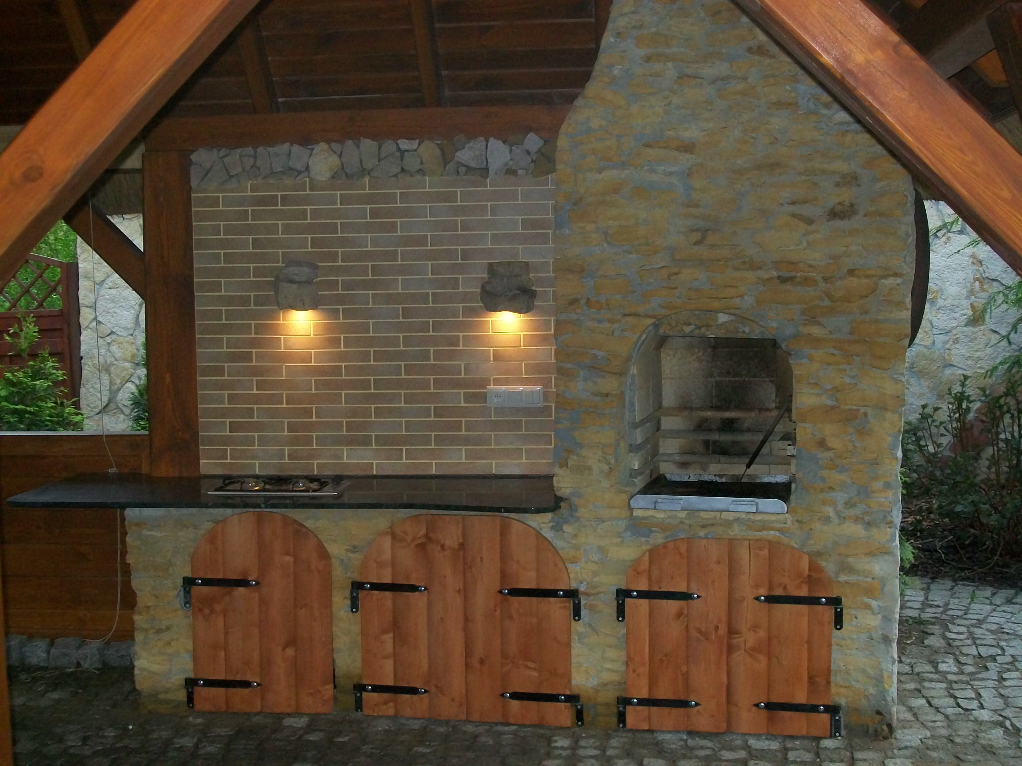 Zadaszona, murowana kuchnia ogrodowa z grillem, blatem roboczym i drewnianymi drzwiczkami, oświetlona dwoma kamiennymi kinkietami.