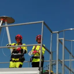 Dwie osoby w kaskach i strojach ochronnych, na platformie z metalową barierką, w trakcie zakładania rękawic ochronnych na tle błękitnego nieba.