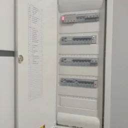 Otwarta biała skrzynka rozdzielcza z listwą zaciskową i wykazem obwodów, widoczna naklejka firmy elektrycznej.