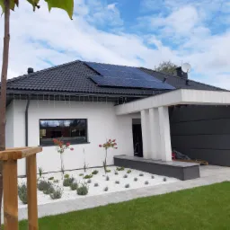 Nowoczesny dom jednorodzinny z czarnym dachem i panelami słonecznymi, widok z zewnątrz na zadbany ogród z białym żwirem.