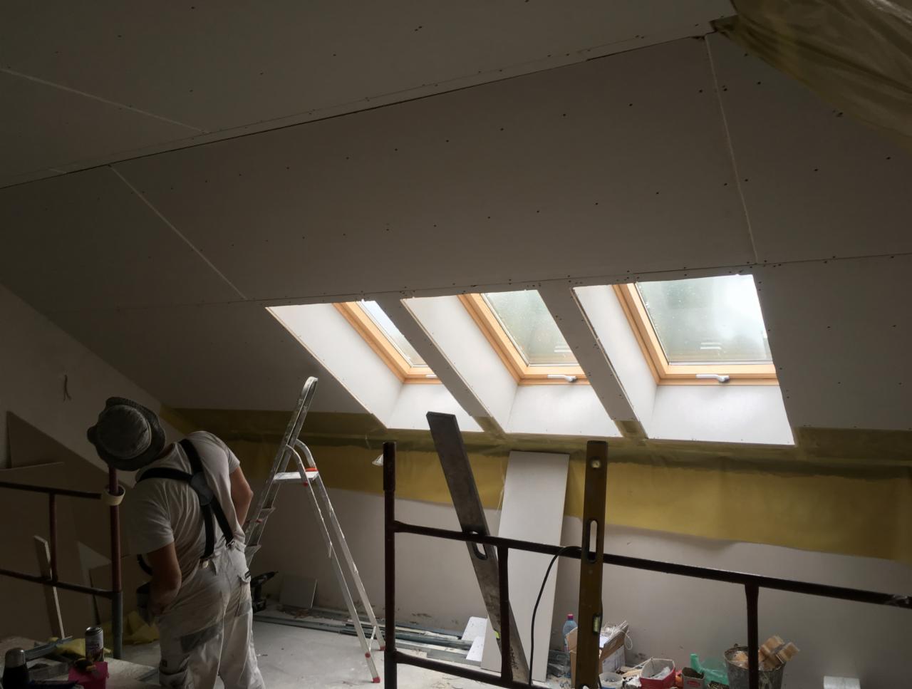 Montaż płyt na poddaszu z oknami dachowymi, widoczny pracownik w stroju roboczym, drabina i narzędzia, żółta warstwa izolacyjna pod płytami.
