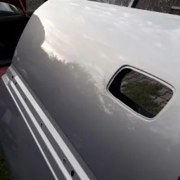 Błyszcząca, srebrna klapa bagażnika samochodowego po lakierowaniu, z widocznym otworem na zamek, leżąca na trawie obok otwartego bagażnika czerwonego auta. Widoczne odbicie nieba i drzew...