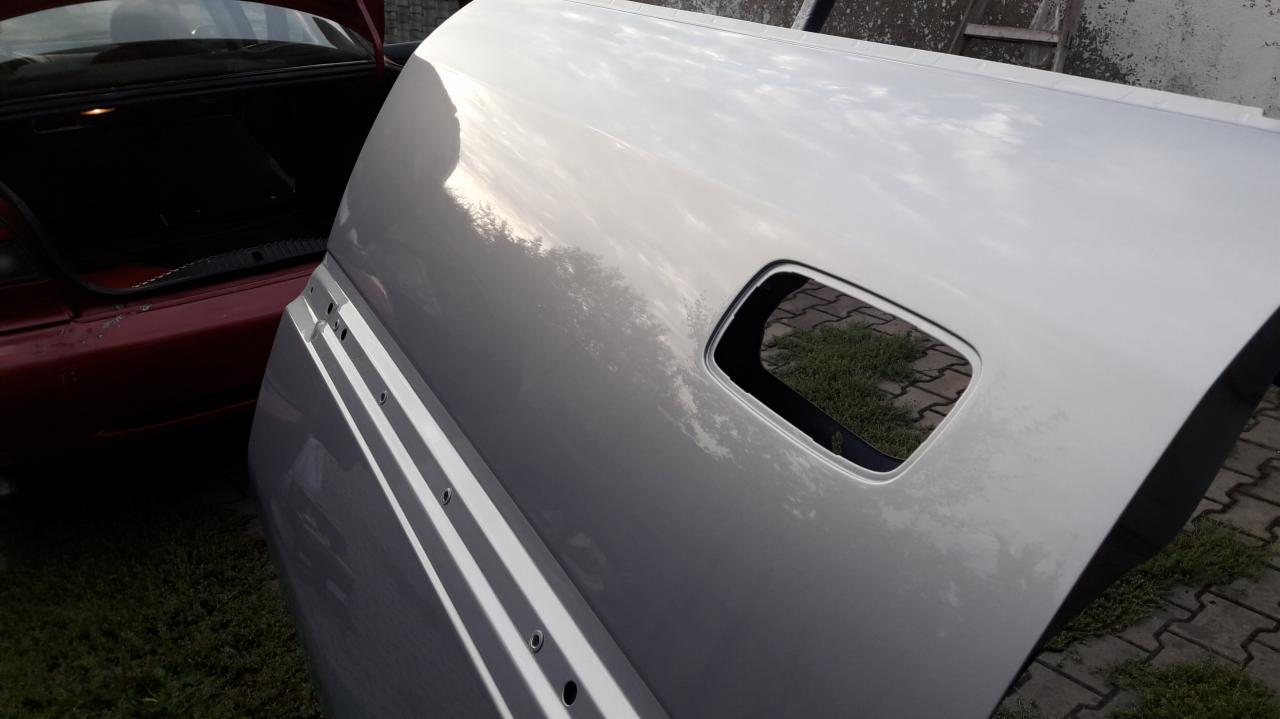 Błyszcząca, srebrna klapa bagażnika samochodowego po lakierowaniu, z widocznym otworem na zamek, leżąca na trawie obok otwartego bagażnika czerwonego auta. Widoczne odbicie nieba i drzew...