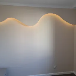 Ściana z dekoracyjnym, podświetlanym wycięciem w kształcie fali, kremowe ściany, biały sufit z listwą ozdobną, szara komoda w rogu.