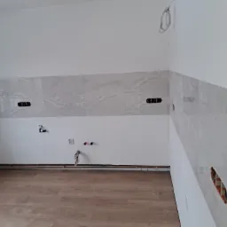Fragment kuchni w trakcie prac wykończeniowych: ułożone płytki imitujące marmur na ścianie, widoczne puszki elektryczne bez osprzętu, wystające rury instalacji wodnej, panele podłogowe.