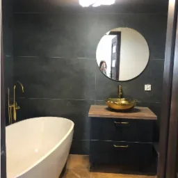 Nowoczesna łazienka z wolnostojącą białą wanną, złotą umywalką nablatową i armaturą, ciemnymi płytkami na ścianach i brązowymi płytkami na podłodze. W lustrze odbija się osoba robiąca zdjęcie.