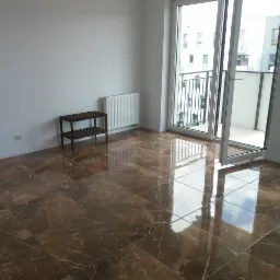 Nowe, błyszczące brązowe płytki podłogowe z marmurkowym wzorem w pustym pokoju z białymi ścianami, grzejnikiem i otwartym balkonem.