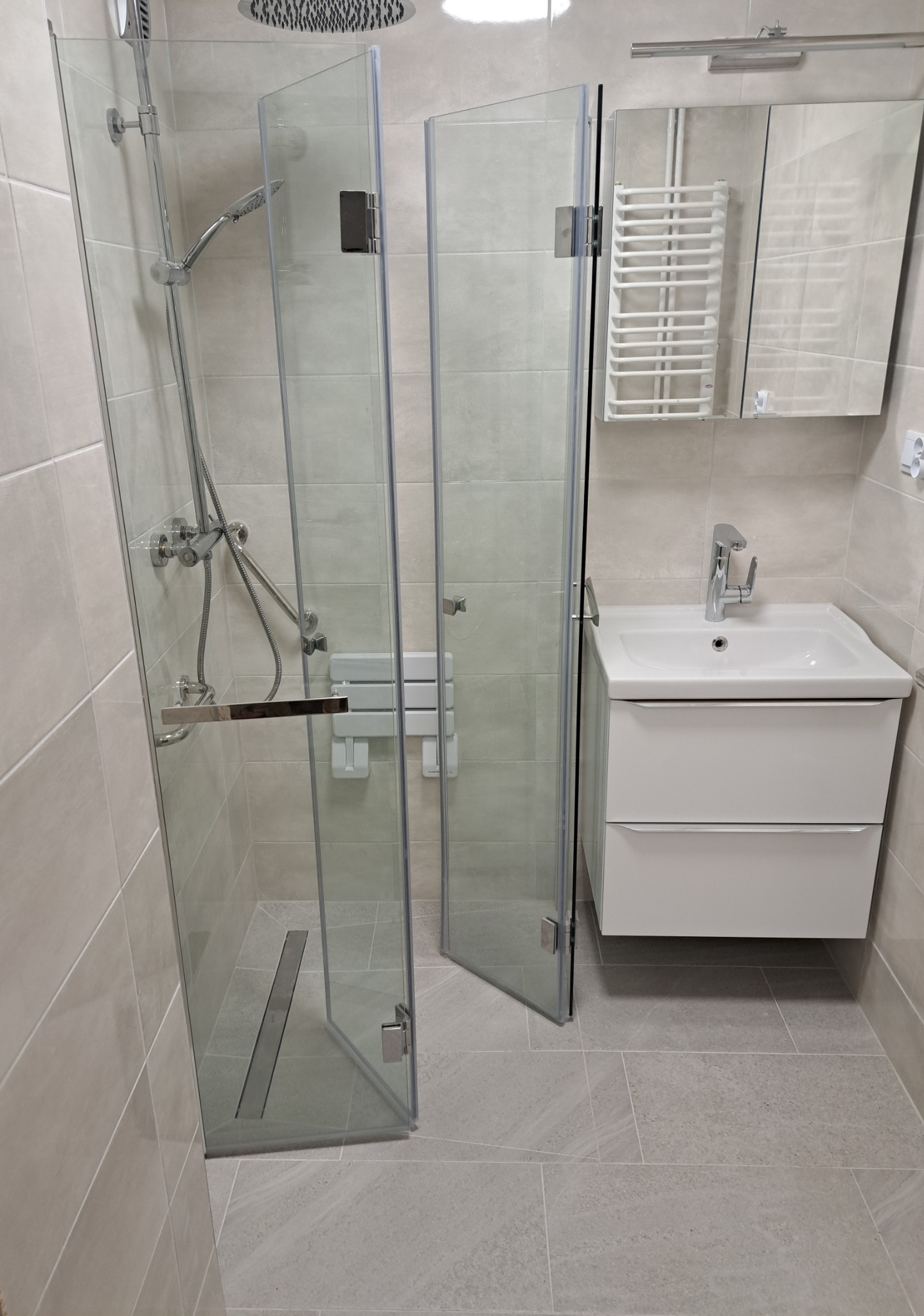 Nowoczesna kabina prysznicowa z siedziskiem i odpływem liniowym, umywalka z szafką i lustrem w odcieniach beżu.