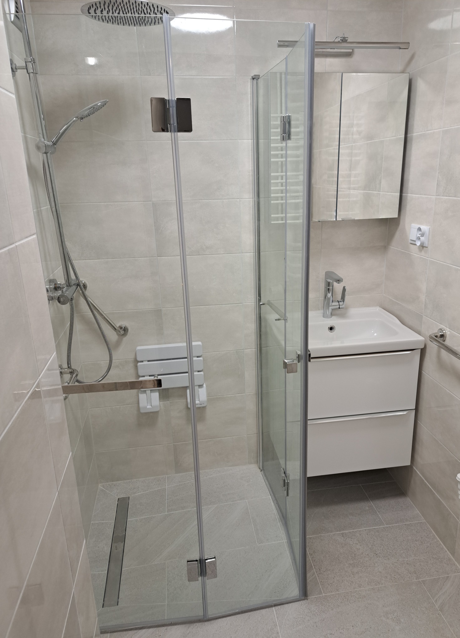 Nowoczesna kabina prysznicowa ze składanym siedziskiem, deszczownicą i odpływem liniowym, umywalka z szafką w jasnej łazience wykończonej płytkami w odcieniach beżu i szarości.