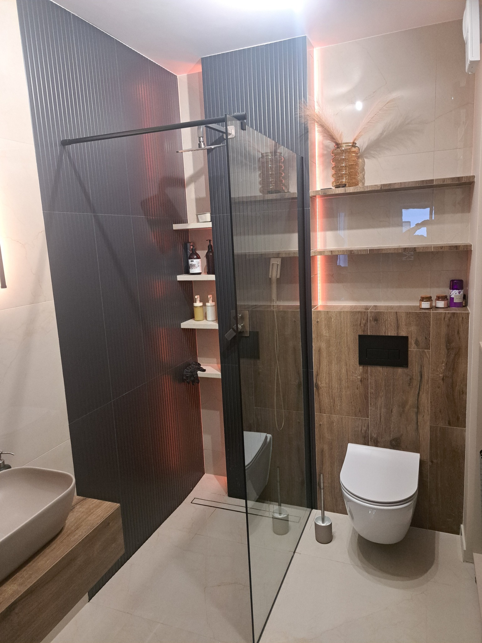 Nowoczesna łazienka z kabiną prysznicową typu walk-in, umywalką nablatową na drewnianej szafce, podwieszaną toaletą i oświetleniem LED akcentującym półki.