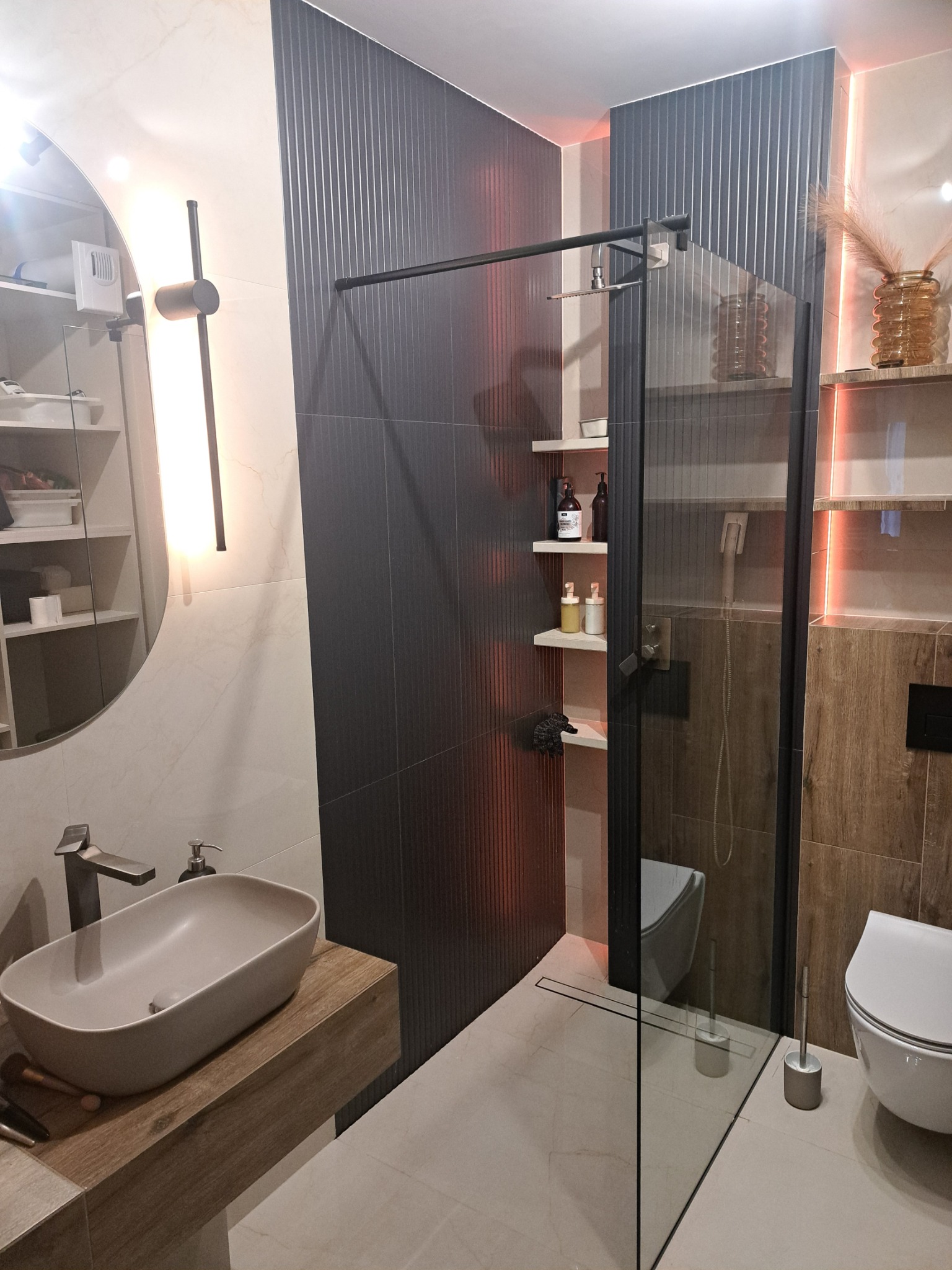 Nowoczesna łazienka z umywalką nablatową, kabiną prysznicową typu walk-in z czarnymi profilami i płytkami imitującymi drewno oraz pionowymi panelami ściennymi w kolorze grafitowym.