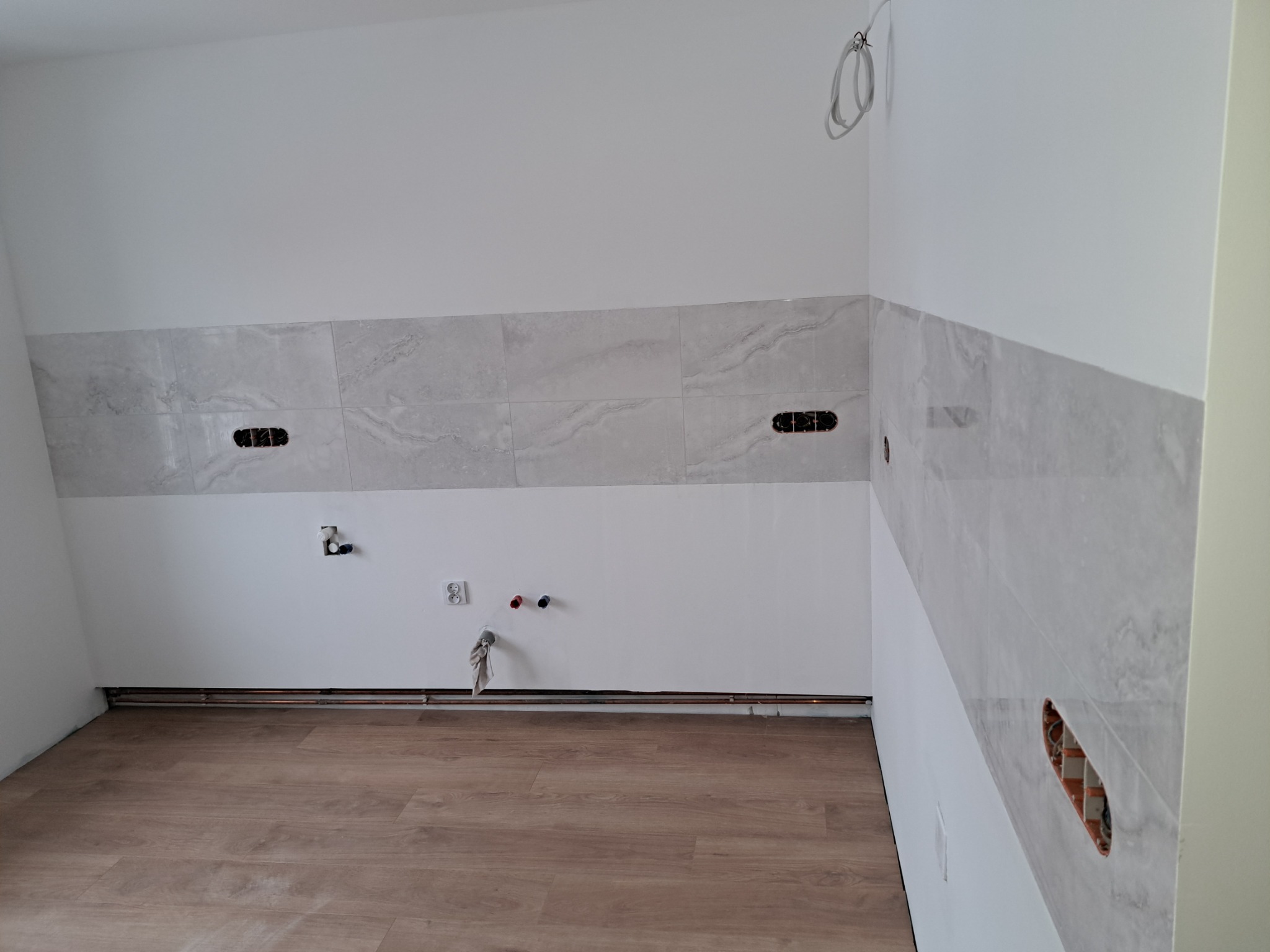 Fragment kuchni w trakcie prac wykończeniowych: ułożone płytki imitujące marmur na ścianie, widoczne puszki elektryczne bez osprzętu, wystające rury instalacji wodnej, panele podłogowe.