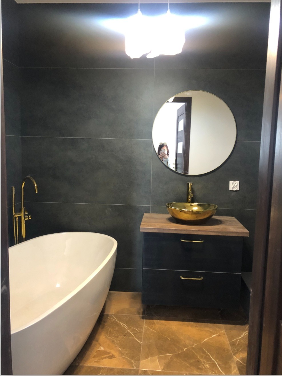 Nowoczesna łazienka z wolnostojącą białą wanną, złotą umywalką nablatową i armaturą, ciemnymi płytkami na ścianach i brązowymi płytkami na podłodze. W lustrze odbija się osoba robiąca zdjęcie.