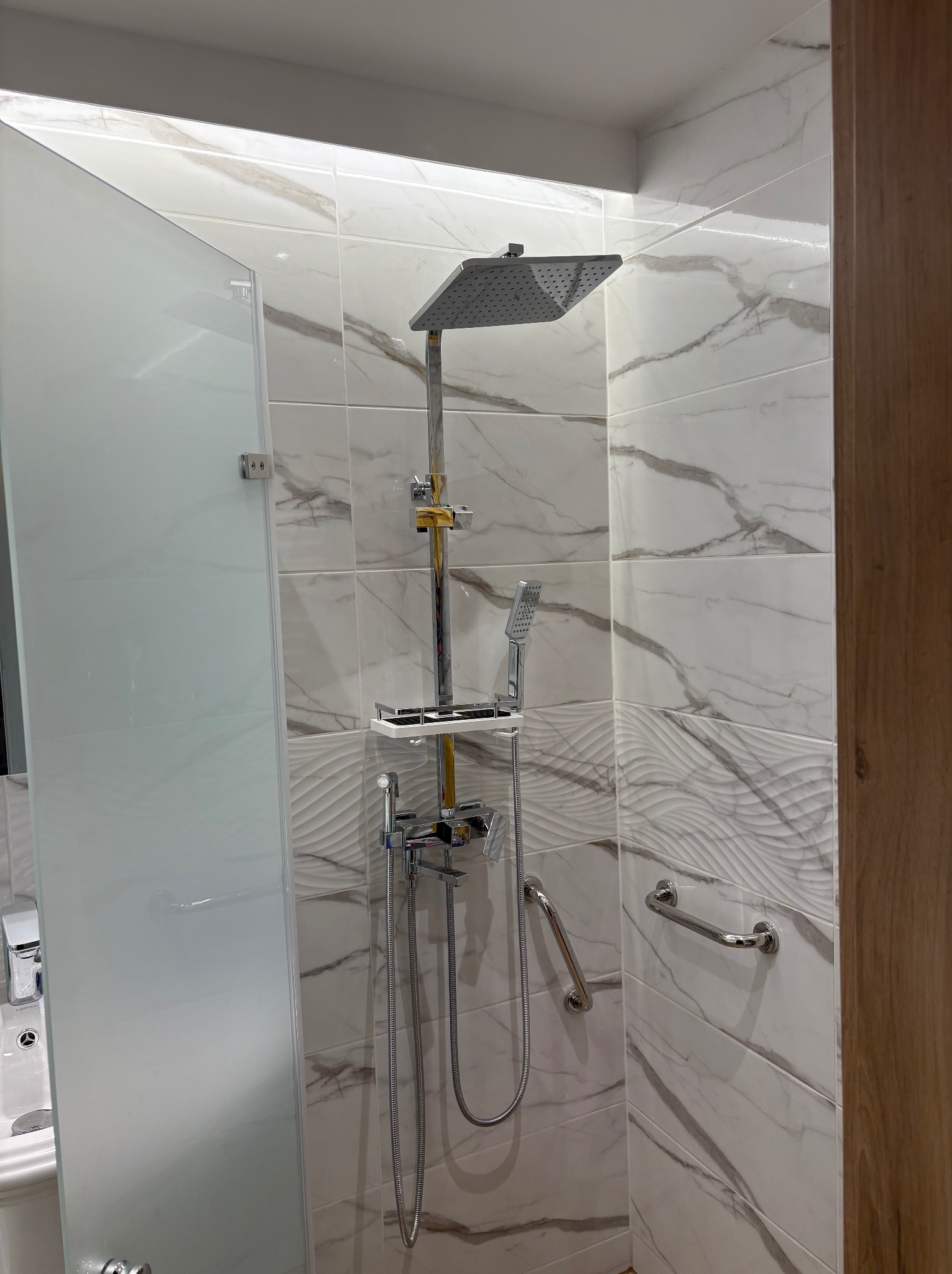 Nowoczesna kabina prysznicowa z deszczownicą i marmurowymi płytkami. Chromowana armatura, szklana ścianka, uchwyt dla niepełnosprawnych. Eleganckie wykończenie łazienki.